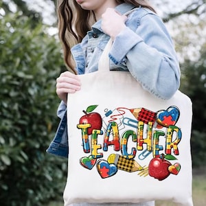 May include: A white tote bag with the word "TEACHER" in colorful letters and school-themed graphics. The design includes red apples, puzzle piece hearts, pencils, and paperclips. The bag is made of a sturdy fabric.