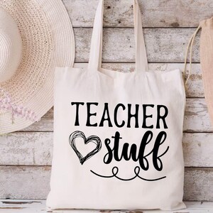 May include: Cream-colored tote bag with the words "TEACHER stuff" in black script and a heart design. A wide-brimmed straw hat, a vase of flowers, and a drawstring bag are also in the image. The background is a rustic, white-washed wooden wall.