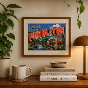 May include: A framed vintage-style postcard print with the words "Greetings from Pendleton." The artwork depicts a scenic view of a town with a waterfall, bridge, and colorful buildings. The print is displayed on a wooden surface with a mug, books, and a lamp.
