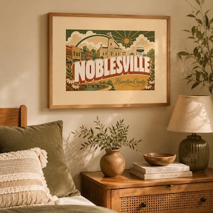 May include: A framed print featuring the word "Noblesville" in large letters, with a vintage-style illustration of a town scene. The artwork includes a courthouse, water tower, and a bridge, with the text "Hamilton County" below. The frame is a light brown color.