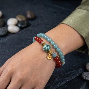 May include: Two beaded bracelets on a wrist. One bracelet features light blue beads, while the other has red beads. A decorative charm with a floral design and gold accents adorns the bracelets. The background includes smooth, gray stones.