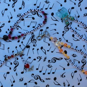 May include: Four beaded lanyards with phrases spelled out in white letter beads. The lanyards are in various colors, including red, pink, orange, and blue. The background is a white fabric with black musical notes.