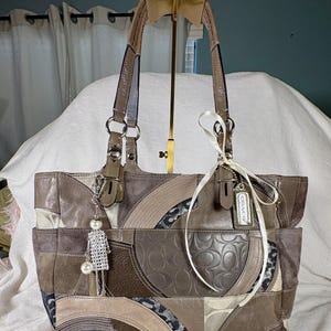 May include: A patchwork leather tote bag in shades of brown, tan, and gray. The bag features a variety of textures and patterns, including suede and embossed leather. It has two handles and a decorative charm. A Coach tag is attached.