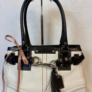 May include: A white leather handbag with black leather trim, dual handles, and a silver-tone metal clasp closure. The bag features a braided black leather strap detail across the front, a hanging black leather tassel, and a light pink ribbon tied to the side.
