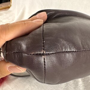 May include: A dark brown leather handbag featuring a smooth, rounded exterior surface with visible stitched seams running along the side. The leather has a subtle sheen and a soft, pliable texture, with a small portion of a red fuzzy accessory visible.