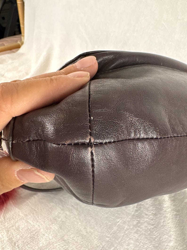 May include: A dark brown leather handbag featuring a smooth, rounded exterior surface with visible stitched seams running along the side. The leather has a subtle sheen and a soft, pliable texture, with a small portion of a red fuzzy accessory visible.