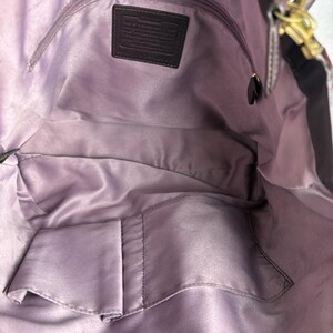 May include: The interior of a dark purple handbag featuring a smooth, light purple satin lining. It includes a zippered pocket with a rectangular leather patch embossed with text, along with two open slip pockets at the bottom for organization.