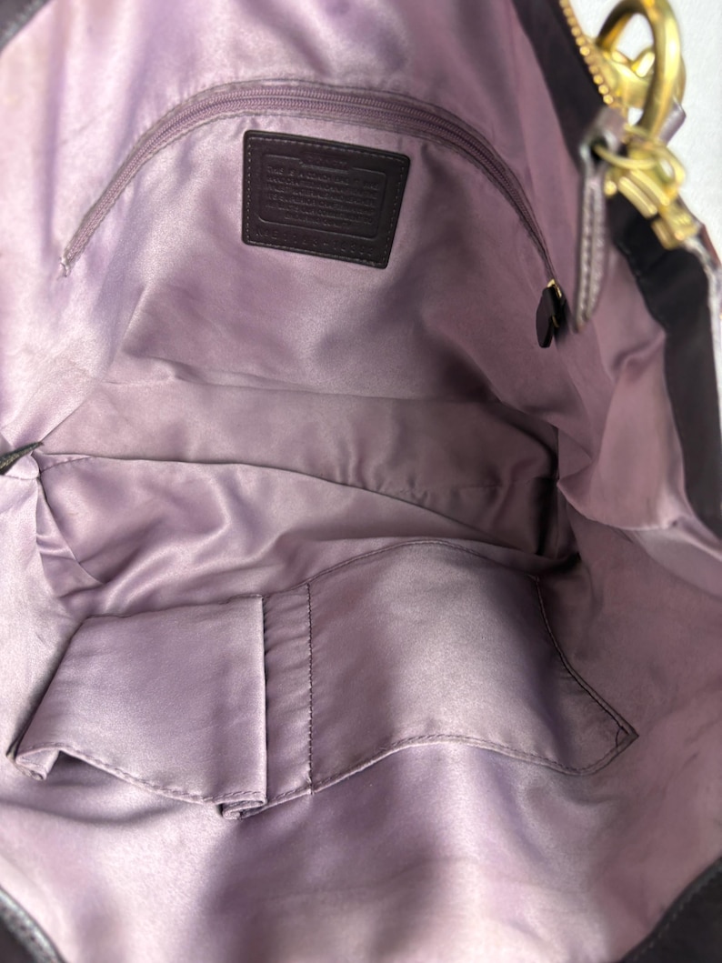 May include: The interior of a dark purple handbag featuring a smooth, light purple satin lining. It includes a zippered pocket with a rectangular leather patch embossed with text, along with two open slip pockets at the bottom for organization.