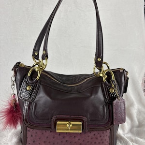 May include: A dark plum leather handbag featuring a textured ostrich-embossed lower panel, gold-tone hardware, a front turn-lock closure with the Coach logo, two shoulder straps, and a decorative dangling charm with a dark pink feather accent.