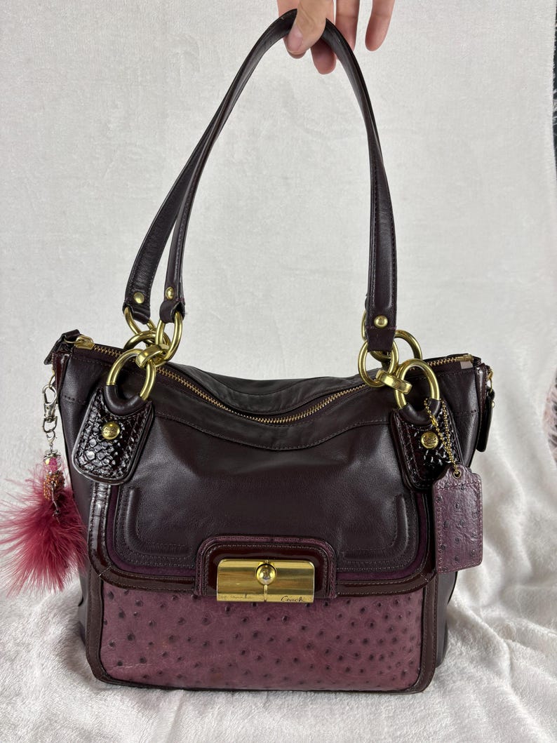 May include: A dark plum leather handbag featuring a textured ostrich-embossed lower panel, gold-tone hardware, a front turn-lock closure with the Coach logo, two shoulder straps, and a decorative dangling charm with a dark pink feather accent.