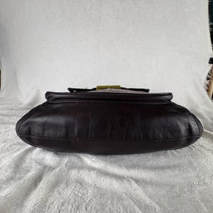 May include: A dark brown leather handbag featuring a smooth, rounded base and a structured top flap. The bag is secured with a rectangular, metallic gold-toned clasp centered on the upper edge, providing a distinct contrast to the dark leather.