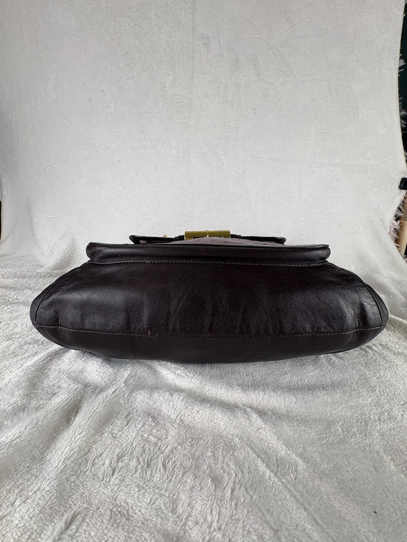 May include: A dark brown leather handbag featuring a smooth, rounded base and a structured top flap. The bag is secured with a rectangular, metallic gold-toned clasp centered on the upper edge, providing a distinct contrast to the dark leather.