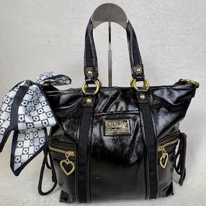 May include: A black patent leather tote bag featuring gold-tone hardware, two top handles, and two front zippered pockets with heart-shaped pulls. A black and white checkered scarf with a floral pattern is tied to the side ring of the bag.