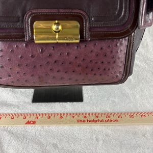 May include: A dark plum leather handbag featuring a textured ostrich-embossed panel, a smooth leather flap, and a gold-tone metal turn-lock closure. A wooden ruler is positioned below the bag, showing a width of approximately 13 inches.