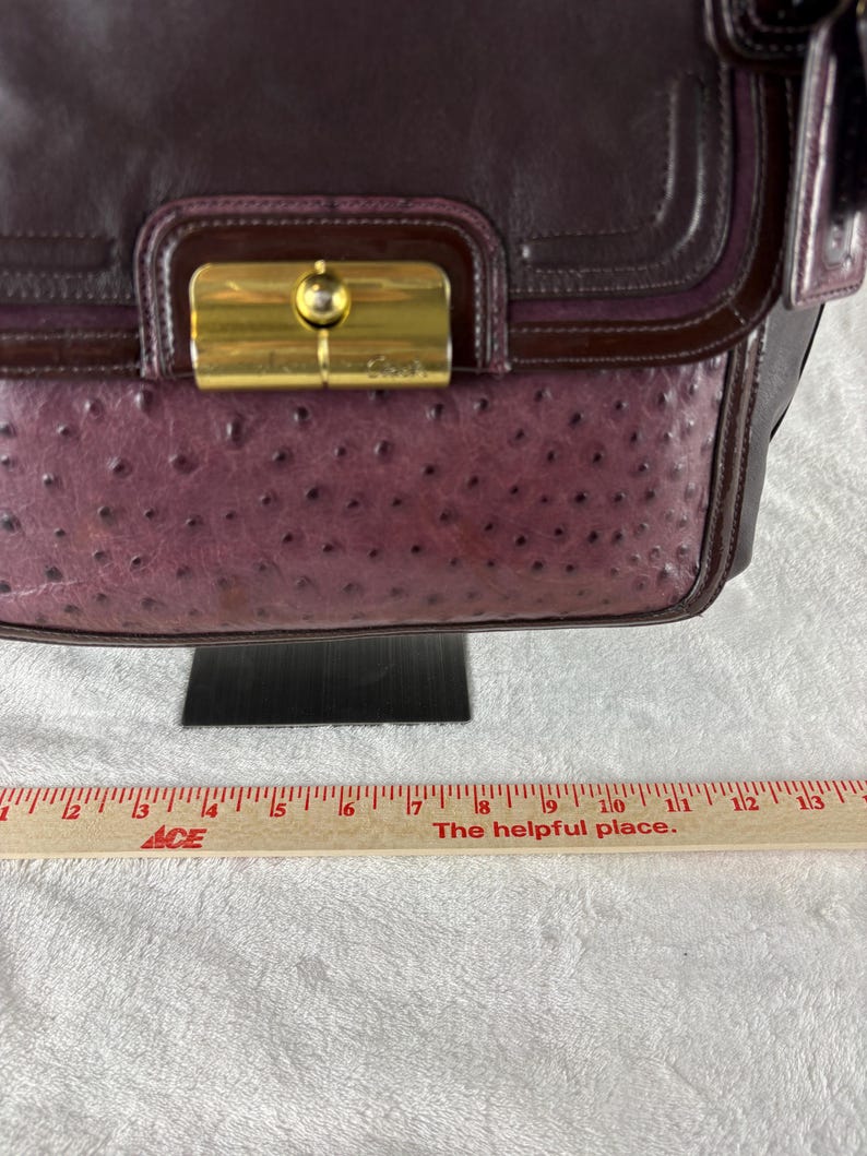 May include: A dark plum leather handbag featuring a textured ostrich-embossed panel, a smooth leather flap, and a gold-tone metal turn-lock closure. A wooden ruler is positioned below the bag, showing a width of approximately 13 inches.