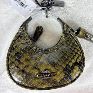 Puede incluir: Un pequeño charm de bolso Coach, verde oliva y negro, con un estampado de piel de serpiente. El charm tiene el logotipo Coach de metal plateado y un llavero a juego. El bolso mide aproximadamente 7,6 cm de ancho.
