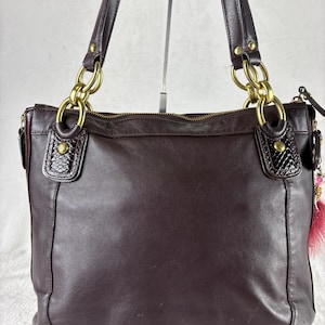 May include: A dark brown leather shoulder bag featuring gold-tone metal hardware, including large chain-link connectors at the base of the two shoulder straps. The bag has textured leather accents at the strap attachments and a zipper closure at the top.