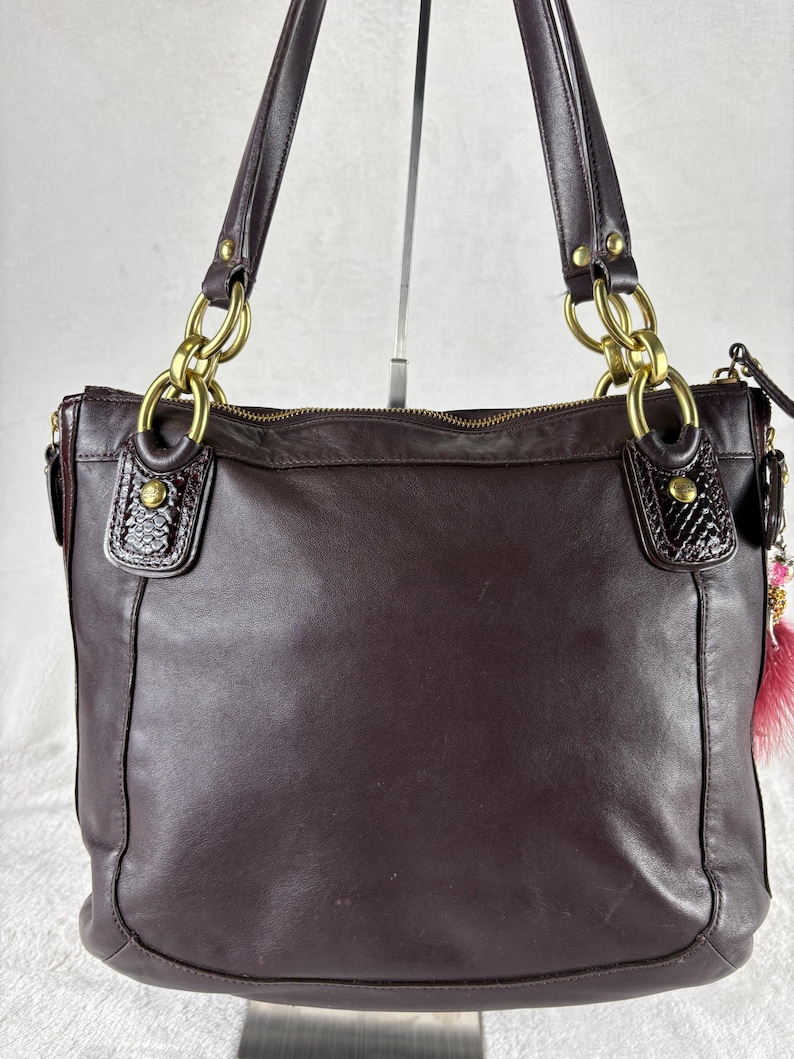 May include: A dark brown leather shoulder bag featuring gold-tone metal hardware, including large chain-link connectors at the base of the two shoulder straps. The bag has textured leather accents at the strap attachments and a zipper closure at the top.
