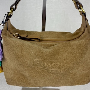 May include: A tan suede handbag featuring a top zipper closure, a brown leather strap with brass hardware, and a multi-colored tassel in lavender, teal, and yellow. The front of the bag is embossed with the Coach logo and text.