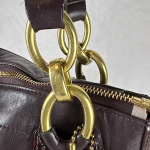 May include: A dark plum leather handbag featuring a gold-tone metal chain link strap attachment. The bag includes a top zipper closure with a gold-tone pull and a small gold-tone ball chain tag hanging from a large circular metal ring connector.