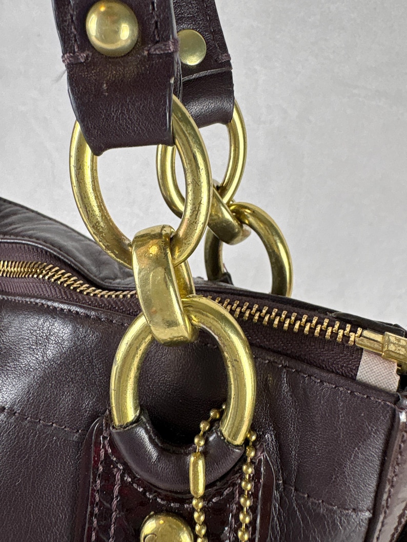 May include: A dark plum leather handbag featuring a gold-tone metal chain link strap attachment. The bag includes a top zipper closure with a gold-tone pull and a small gold-tone ball chain tag hanging from a large circular metal ring connector.