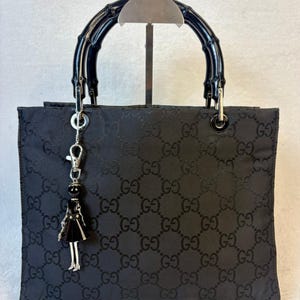 May include: A black Gucci handbag featuring the signature GG monogram pattern, constructed with a structured rectangular body and a dark, polished bamboo handle. A silver-toned metal charm shaped like a small figure hangs from the handle.