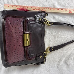 May include: A dark purple leather shoulder bag with a textured ostrich-patterned front panel, gold-tone hardware, and a flap closure. A wooden ruler placed above the bag shows a width of approximately 12 inches for the main body of the handbag.