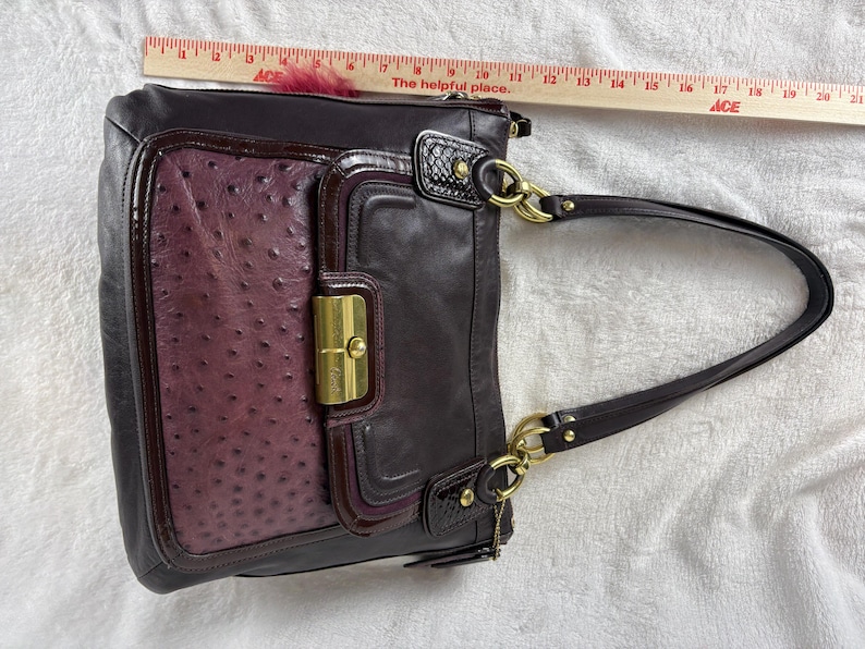 May include: A dark purple leather shoulder bag with a textured ostrich-patterned front panel, gold-tone hardware, and a flap closure. A wooden ruler placed above the bag shows a width of approximately 12 inches for the main body of the handbag.