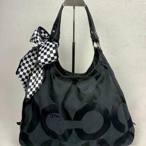 May include: A black hobo bag featuring a tonal circular pattern and the Coach logo. The bag has black leather handles and a black and white checkered ribbon tied to one side. The hardware is silver-toned, and the bag has a slouchy silhouette.
