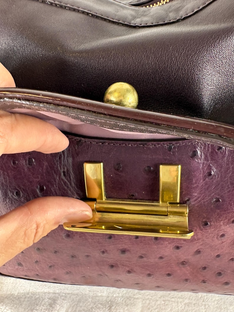 May include: A dark purple handbag featuring a textured ostrich-style leather finish. The bag includes a gold-toned metal flip-lock closure mechanism and a small gold-toned spherical knob accent on the top edge, with a light purple interior lining.