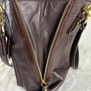 May include: A dark purple leather handbag featuring a prominent diagonal gold-tone metal zipper closure on the front panel. The bag includes gold-tone hardware, a leather zipper pull, and dark brown leather trim accents along the zipper edges.
