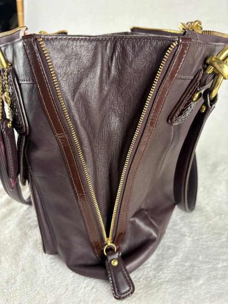 May include: A dark purple leather handbag featuring a prominent diagonal gold-tone metal zipper closure on the front panel. The bag includes gold-tone hardware, a leather zipper pull, and dark brown leather trim accents along the zipper edges.