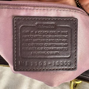 May include: A dark brown leather rectangular patch sewn onto a light purple fabric lining inside a bag. The patch features the embossed Coach logo and text stating it was handcrafted in China with leather, including the serial number 1193-18303.