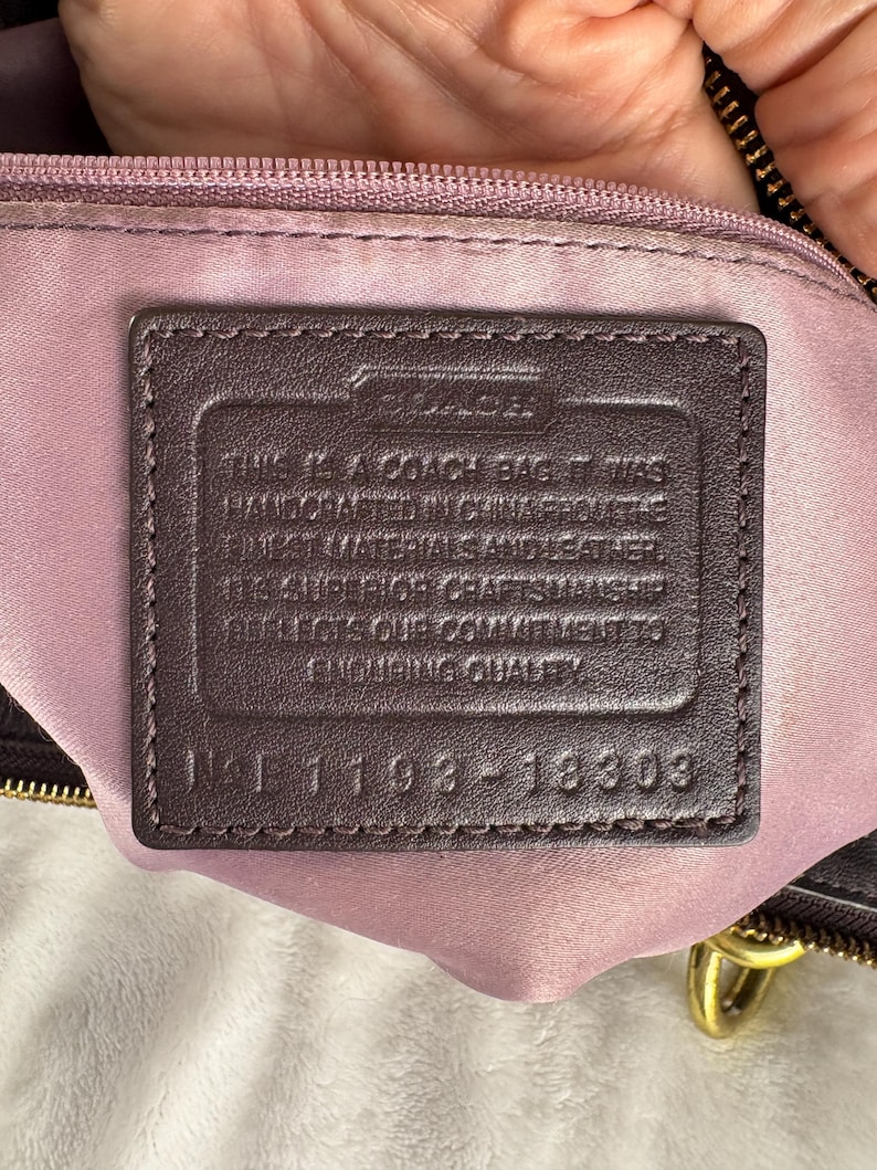 May include: A dark brown leather rectangular patch sewn onto a light purple fabric lining inside a bag. The patch features the embossed Coach logo and text stating it was handcrafted in China with leather, including the serial number 1193-18303.