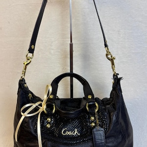May include: A black leather handbag featuring a textured snakeskin-patterned panel, gold-tone hardware, dual top handles, and a detachable shoulder strap. The front displays the gold Coach logo, a hanging leather tag, and a decorative cream ribbon.