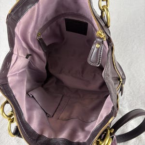 May include: A dark purple leather handbag with a light purple fabric interior lining. The bag features gold-tone metal hardware, including large chain-link handles, a top zipper closure, and an interior zippered pocket with a leather brand patch.