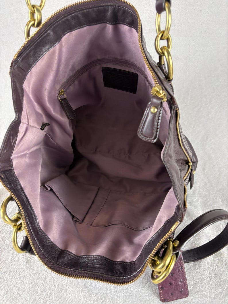 May include: A dark purple leather handbag with a light purple fabric interior lining. The bag features gold-tone metal hardware, including large chain-link handles, a top zipper closure, and an interior zippered pocket with a leather brand patch.