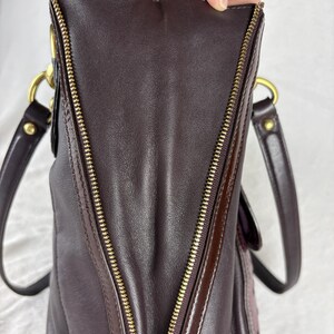 May include: A dark brown leather handbag featuring a prominent gold-tone metal zipper running vertically along the side panel. The bag includes two dark brown leather shoulder straps attached with gold-tone metal hardware rings and rivets.