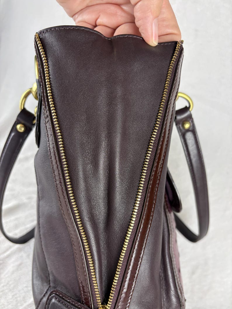 May include: A dark brown leather handbag featuring a prominent gold-tone metal zipper running vertically along the side panel. The bag includes two dark brown leather shoulder straps attached with gold-tone metal hardware rings and rivets.