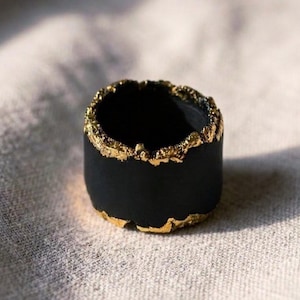 May include: A wide, cylindrical ring featuring a matte black finish on the exterior and interior. The top and bottom edges of the band are accented with a textured, metallic gold leaf application that creates an irregular, organic appearance.