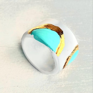 May include: A white ring with a square shape, featuring turquoise, gold, and brown accents. The ring has a modern design with a bold color palette. The ring is a fashion accessory.