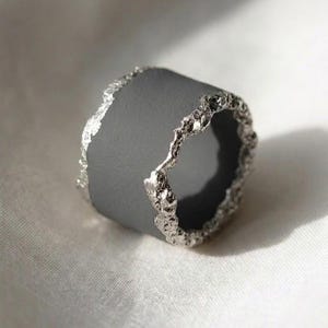 May include: A wide band ring featuring a smooth matte charcoal gray center section contrasted by two irregular, textured silver-toned edges with a rough, organic finish that frames the dark band on both sides.