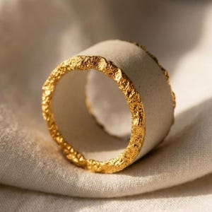 May include: A wide band ring featuring a smooth, matte off-white center section contrasted by two textured, metallic gold edges. The ring has a cylindrical shape with a consistent width throughout, showcasing a distinct combination of materials.