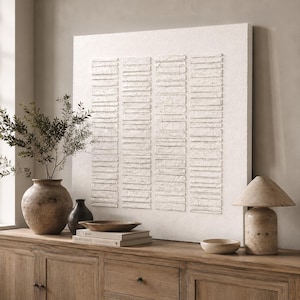 May include: A square white wall art piece featuring four vertical columns of raised, horizontal rectangular segments with a textured finish. The artwork is displayed on a wooden surface alongside a large ceramic vase, a smaller dark vase, a stack of books, a shallow bowl, and a table lamp.