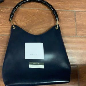 May include: A navy blue handbag with a bamboo handle and gold-colored hardware. The bag has a rectangular white tag with the word "GUCCI" printed on it, and a smaller tag below it. The bag is made of a smooth material.