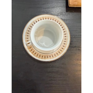 May include: A white teacup and saucer set with a gold rim and a repeating geometric pattern in shades of brown. The cup sits inside the saucer, ready for tea or coffee. The set is on a dark surface.