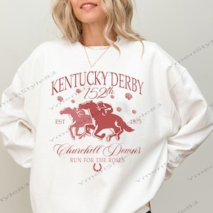 May include: A white sweatshirt with a maroon graphic featuring two racehorses, the text "Kentucky Derby 152th", and "Churchill Downs". The design includes roses and a horseshoe. The sweatshirt is worn by a person.