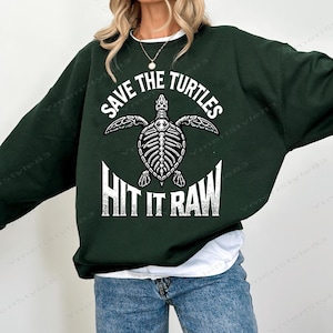 May include: Dark green sweatshirt with the text "SAVE THE TURTLES" above a white skeletal turtle graphic. Below the turtle is the text "HIT IT RAW" in white. The sweatshirt is paired with blue jeans.
