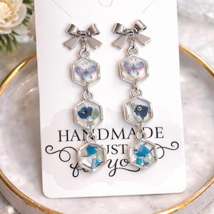 Peut inclure: Boucles d'oreilles pendantes argentées avec un nœud en haut. Chaque boucle d'oreille comporte trois breloques hexagonales avec des motifs floraux et papillons. Le texte "HANDMADE JUST for you" est visible.
