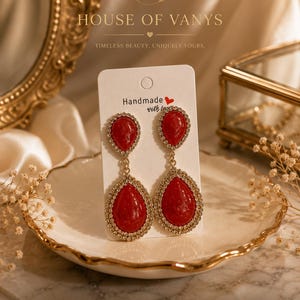 May include: A pair of gold-tone drop earrings featuring two red teardrop-shaped stones each, surrounded by small clear crystals. The earrings are attached to a white display card with the text Handmade with love and a red heart icon.